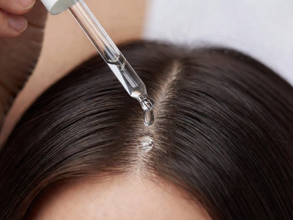 Close-up of a glass pipette dropper applying a clear hydrating serum along a clean scalp parting